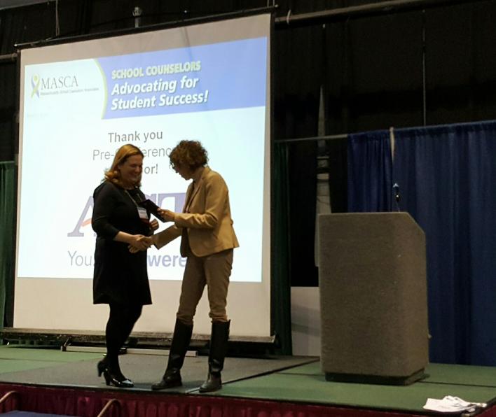 Dr. Laurie Dickstein-Fisher shaking hands with a presenter on stage as she receives her School Counselor of the Year award. An illegible slide is projected in the background. Dr. Dickstein-Fisher and the presenter are standing in front of a podium.