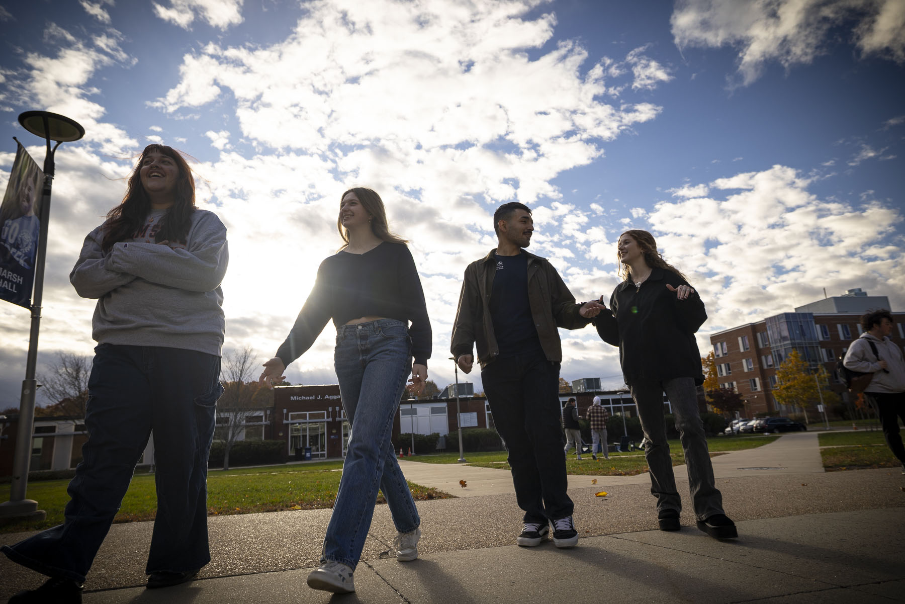 Four Salem State University students walk together and laugh on campus on a sunny fall day, with residence halls visible in the background.
