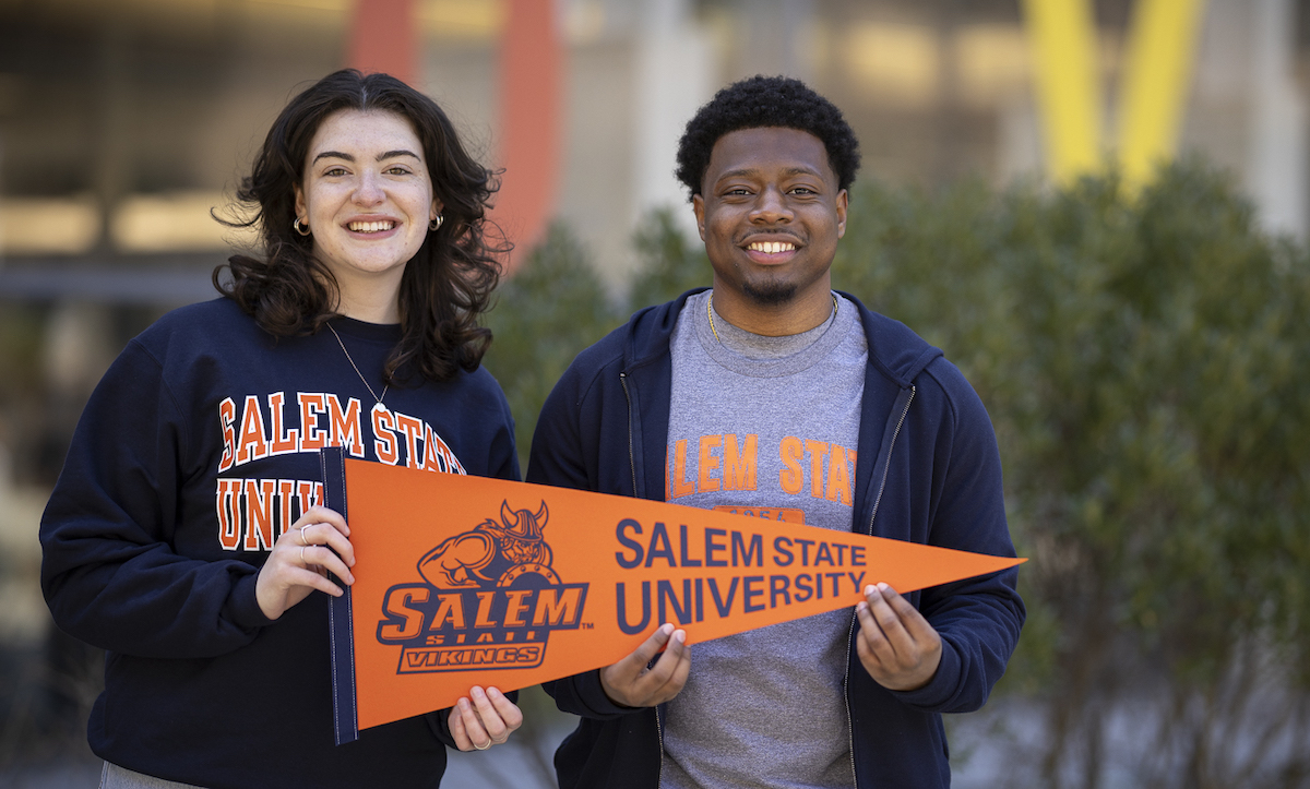 Two undergraduate students smiling in front of the Berry Library with a Salem State Vikings pennant