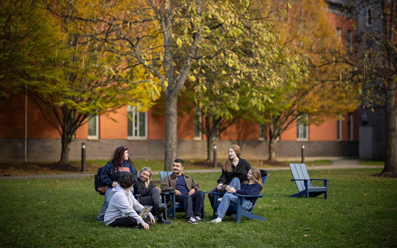 A group of Salem State University students relax and talk together in Adirondack chairs on a grassy quad surrounded by trees with fall foliage.