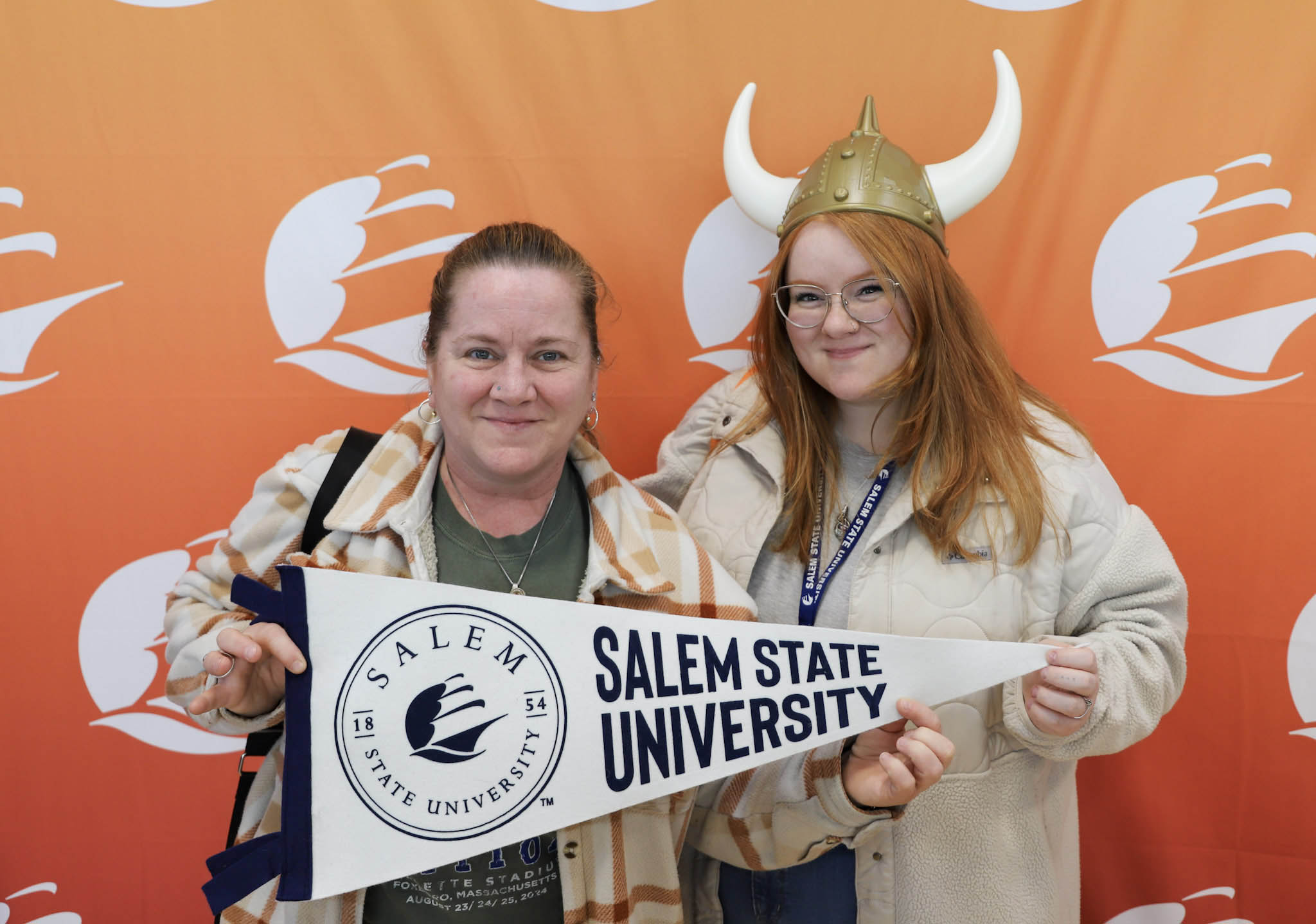 A new student stands with her mom in a photo booth, wearing a Viking helmet with hors, holding a Salem State pennant