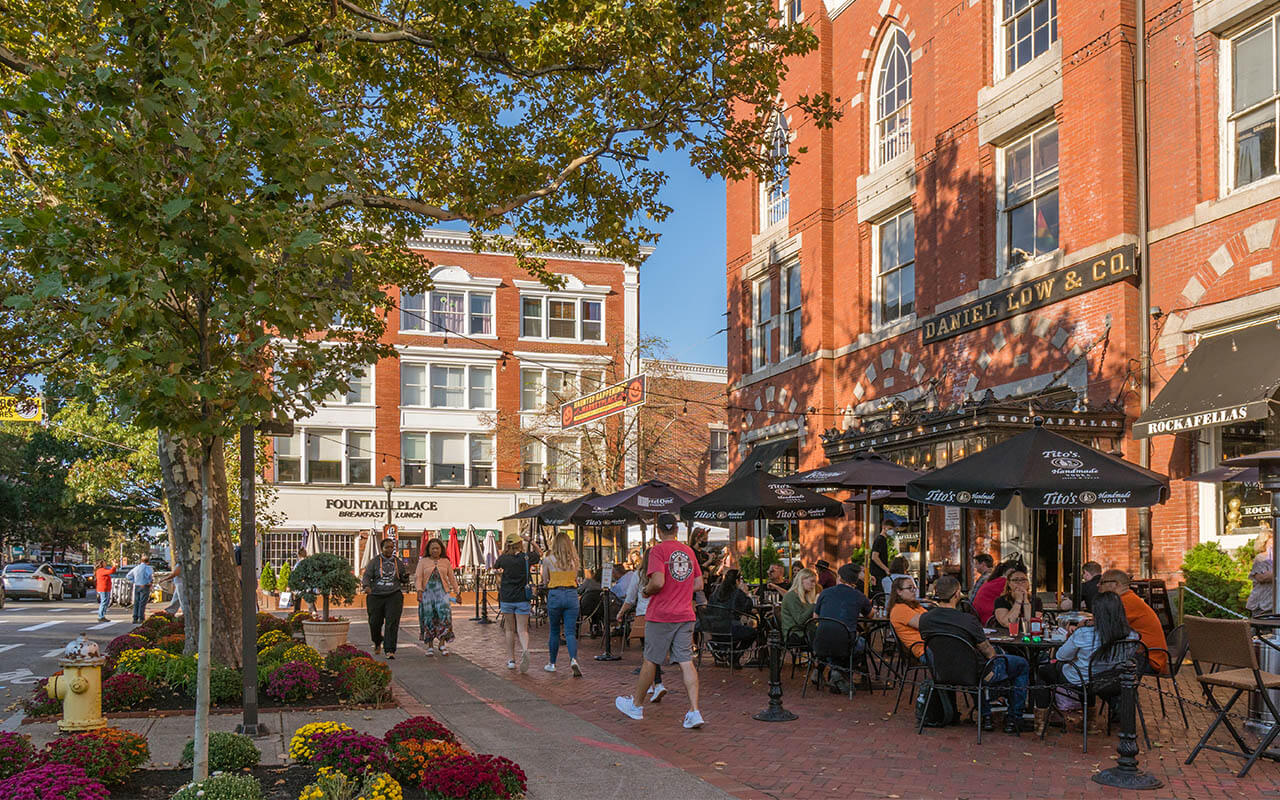 People dining outdoors and walking along a brick-lined sidewalk in downtown Salem, Massachusetts, surrounded by fall flowers and historic red-brick buildings.