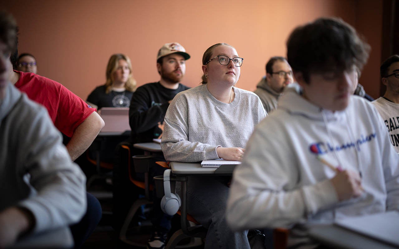 Students seated at desks in a classroom attentively listening during a lecture, with one female student in focus wearing glasses and a gray sweatshirt.