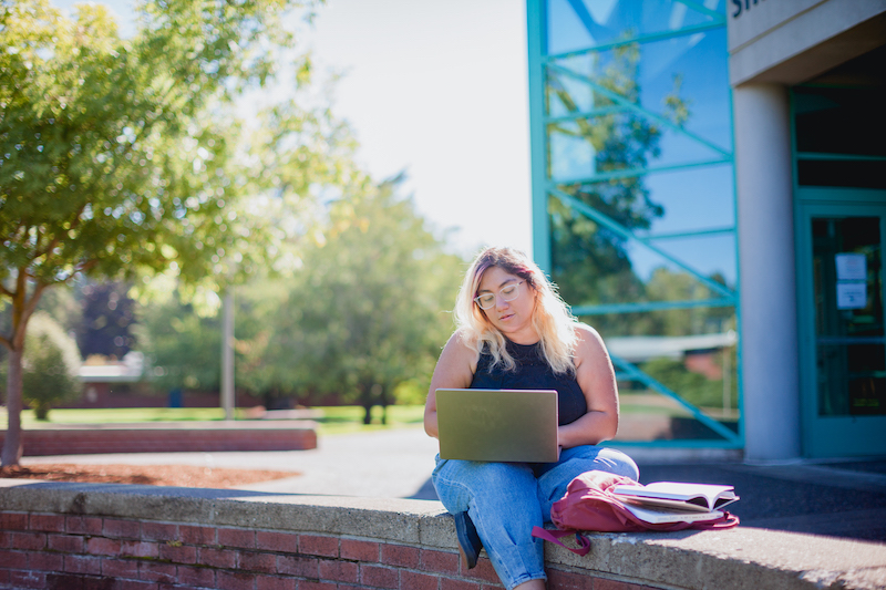 A female student is sitting outside a tall building with a laptop.