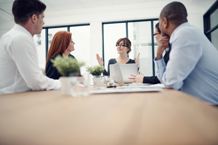 Stock Photo of a female administrator takes the lead talking at table of doctors and other staff.