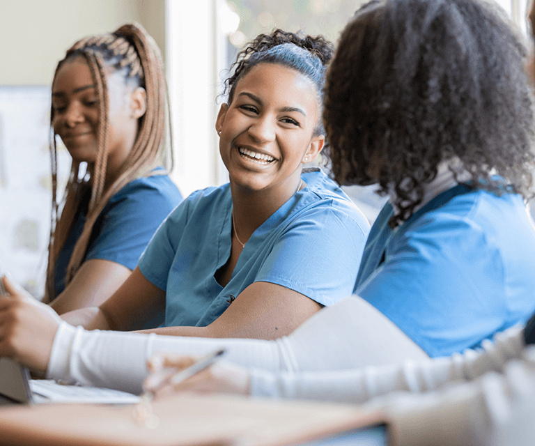 A nursing student wearing scrubs sits with two of her peers and has a conversation.