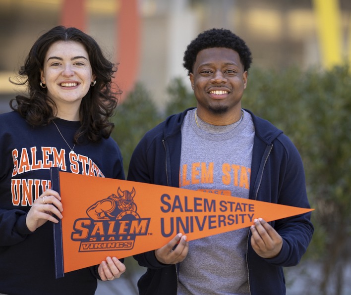 Two undergraduate students smiling in front of the Berry Library with a Salem State Vikings pennant