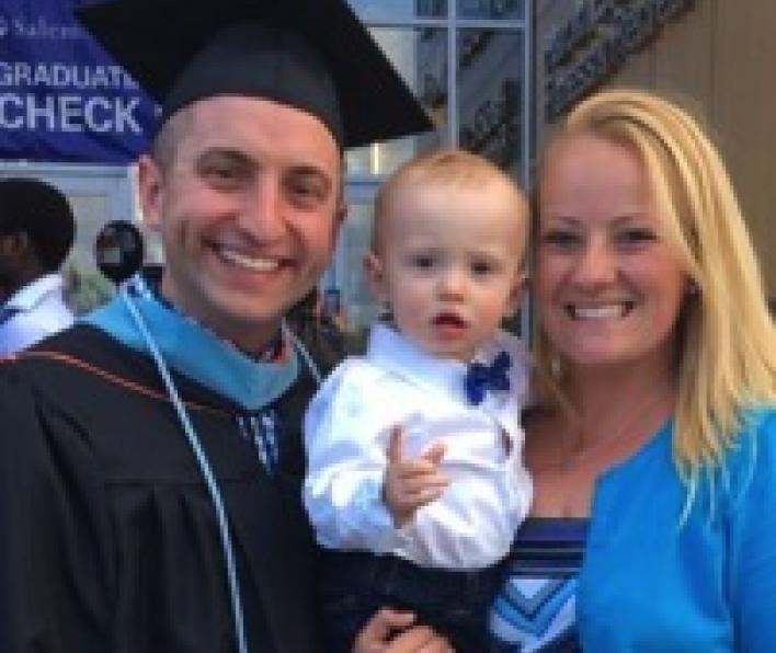 A graduate in cap and gown poses with his wife and young son. The image is of the waist up.