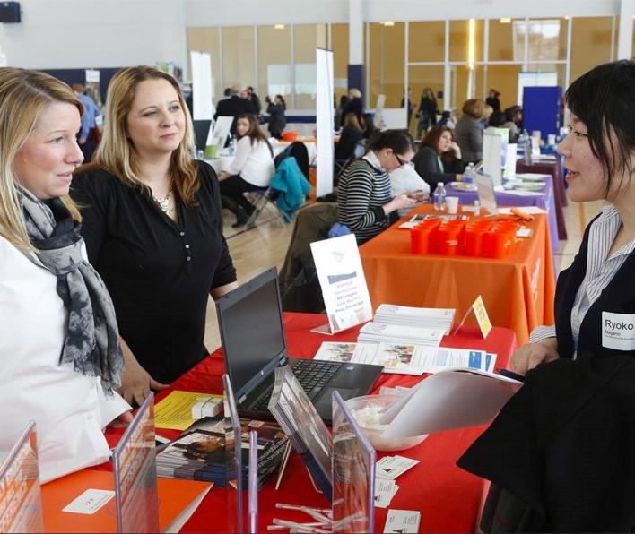 A student in a blazer stands at a table and speaks with two women who are behind the table. They are in an auditorium with other employers at tables.