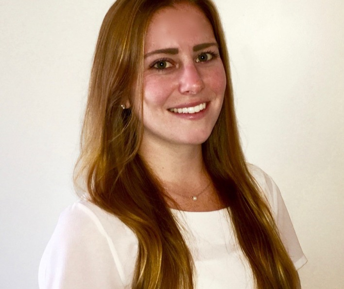 Image of a young adult white woman wearing a white t-shirt. She has long dark-blond hair. She is looking towards the viewer.