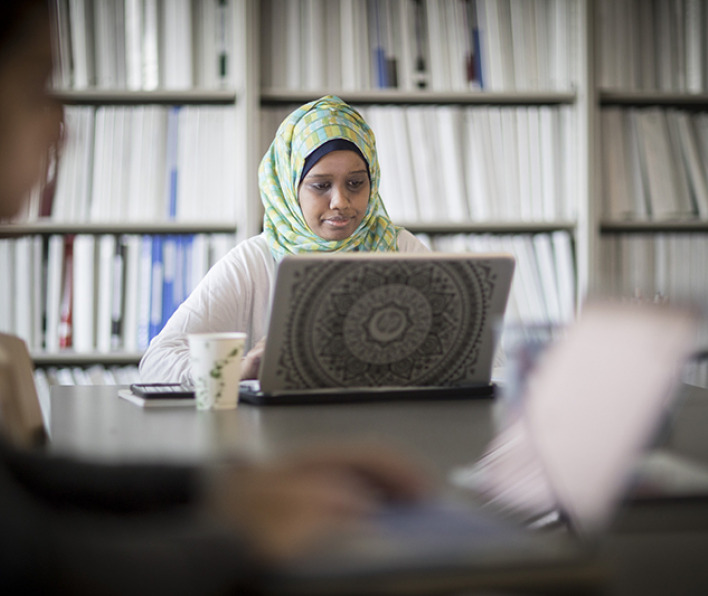 girl wearing headscarf, sitting at a large desk in front of a wall of bookshelves, using her laptop