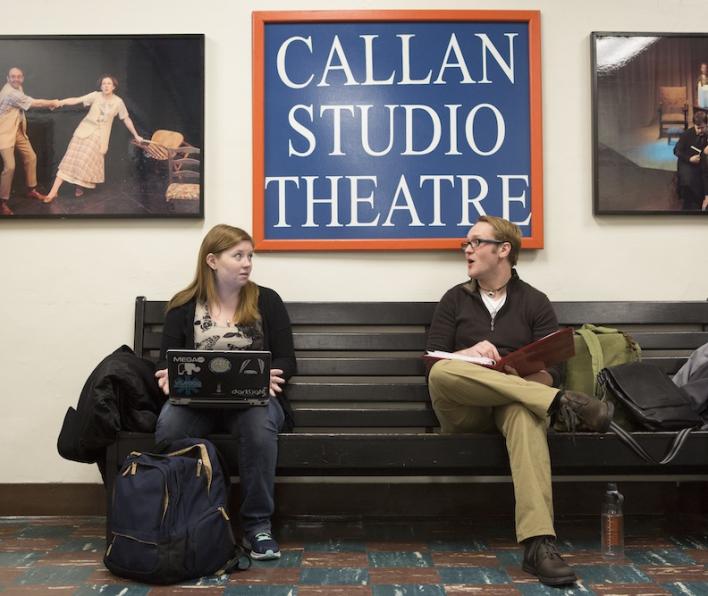 Callan Studio Theatre sign hangs on a wall next to theatre photos as two students sit on their laptops and talk on a bench.