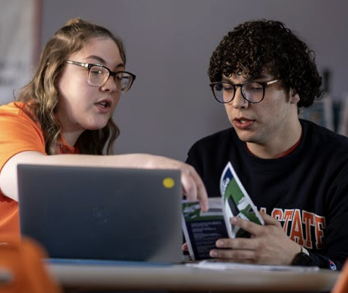 A woman points something out to a man behind a laptop