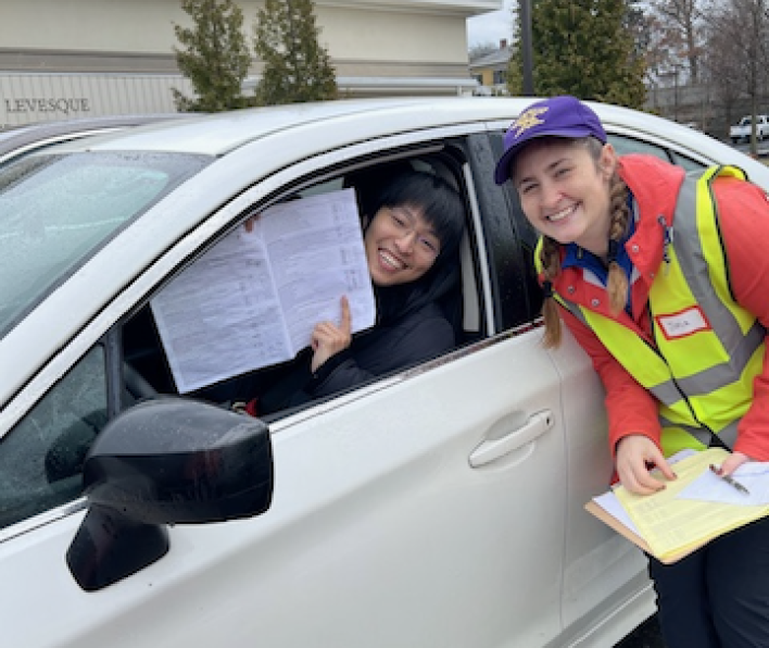 woman in a car holds up paper, woman next to car in a reflective jacket 