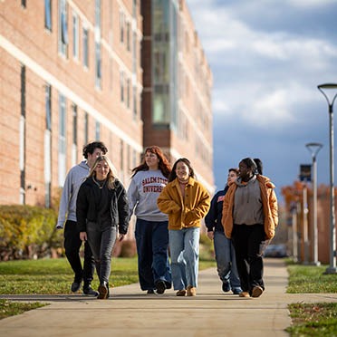 A group of Salem State University students walk together along a campus pathway on a sunny afternoon, talking and wearing fall clothing.