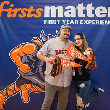 A father and daughter stand in front a backdrop to pose with Salem State University pennants in their hands
