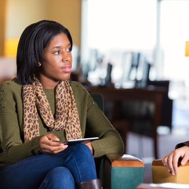 A student in jeans and a sweater sitting in a chair talking to another student