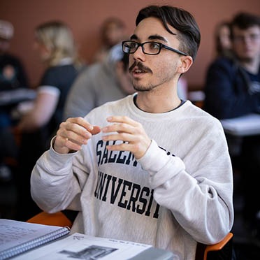 A first-year male student engages in a classroom discussion, enthusiastically moving his hands as he discusses a topic with his classmates and professor