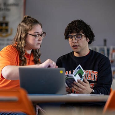 A Salem State student and student staff member sit together at a table, reviewing a booklet and laptop. The student staff member gestures to the screen, offering guidance.