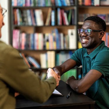 Smiling student shaking hands with another person across a desk, with bookshelves in the background, suggesting a job interview or advising meeting.