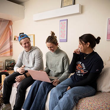A male student and two female students hanging out in a Forten Hall dorm room, looking at coursework on a laptop