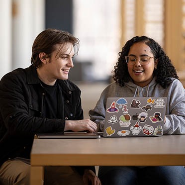 Two Salem State University students sit at a table with a laptop, talking and gesturing as they collaborate in a bright meeting room.