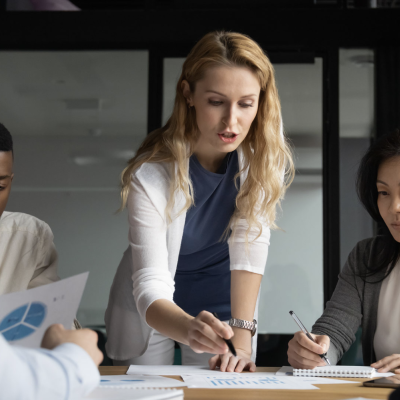 A group of co workers sit at a table and look at graphs. A woman stands and points something out on a piece of paper.