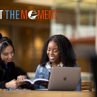 Two students sit at a laptop under the "Meet the Moment" logo