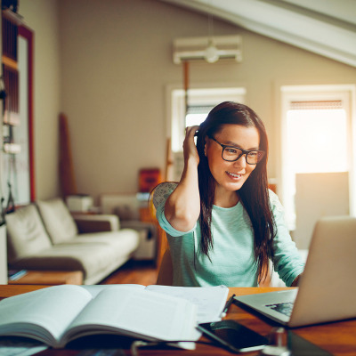 A young adult student sits at a desk in her living room, with a notebook open, smiling at her laptop.