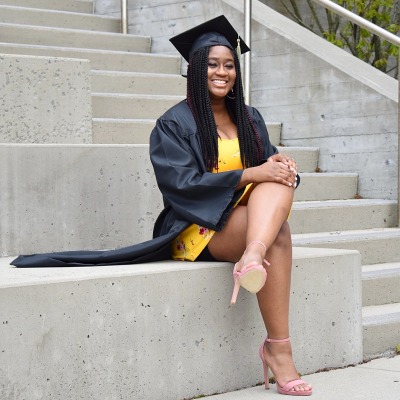 Janique Jean, a 2020-2021 STEP Coach, sits on steps at Salem State in her cap and gown after receiving her undergraduate degree.