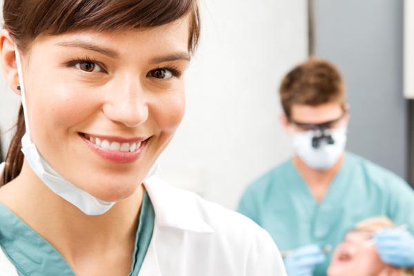 A dental assistant smiles with a dentist and patient in the background