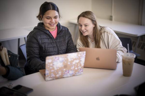Two students sitting at a table with laptops opened, smiling at the screens