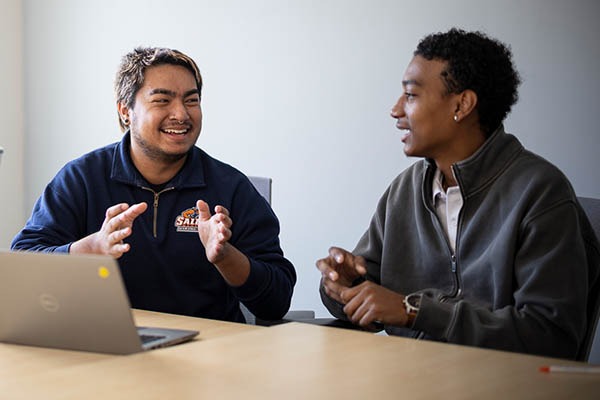 Two Salem State University students sit at a table with a laptop, talking and gesturing as they collaborate in a bright meeting room.