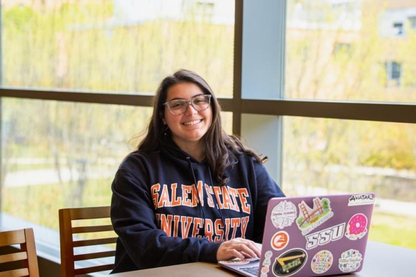 A student smiles from behind a laptop in the library