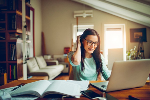 A young adult student sits at a desk in her living room, with a notebook open, smiling at her laptop.