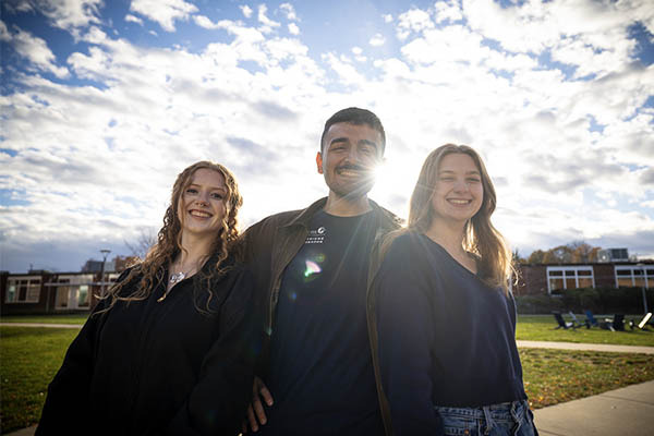 Three Salem State students stand together outdoors on campus, smiling with the sun shining behind them and campus buildings in the background.