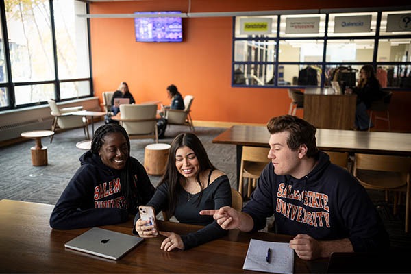 Three Salem State University students sit together in the commuter lounge, smiling and looking at a phone while studying with a laptop and notebook.