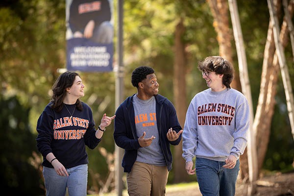Three Salem State University students walk together on a tree-lined path, talking and wearing Salem State sweatshirts.