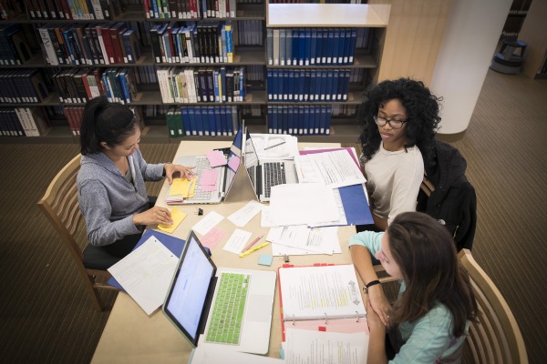 A group of girls sitting together at a table with papers scattered around and laptops sitting in front of them.