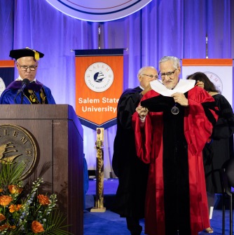 Carlos Santiago ’25H, retired commissioner of the state’s Department of Higher Education, is hooded by Salem State University Trustee Paul Mattera and Trustees board chair Samanda Morales, as Santiago receives his honorary degree at the final commenc