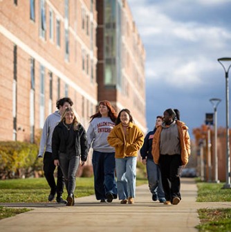 A group of Salem State University students walk together along a campus pathway on a sunny afternoon, talking and wearing fall clothing.