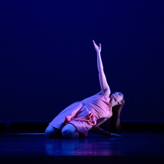 Dancer kneeling on the stage extending right arm upward, surrounded by blue light