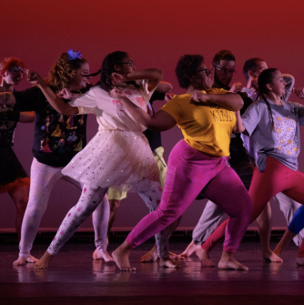 A group of dancers in multi-color outfits with arms raised