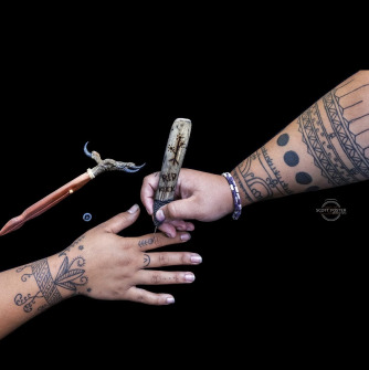 Two hands in front of a black background. One hand is holding a hand poke tattooing device while the other is getting tattooed.