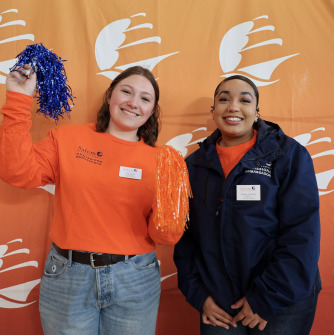Two people stand in front of an orange backdrop with the Salem State logo, one person shakes a blue pom pom