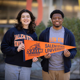 A white female student and Black male student dressed in Salem State apparel stand happily in front of the university library pridefully holding a Salem State pennant 