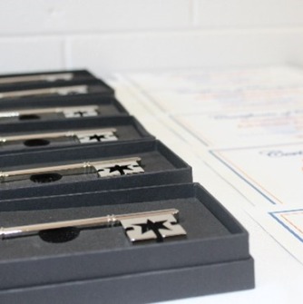 Photo of silver keys in boxes (award that recipients receive) with the corner of a pile of certificates also appearing