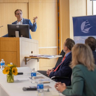 Brian Kelly is standing behind a podium with a computer monitor on it and gesturing. To the right, three individuals are sitting at a table with their backs to the camera; they are turned toward Kelly.