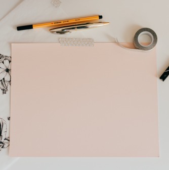 Pieces of light pink paper with scissors, pencils, and pens around it.