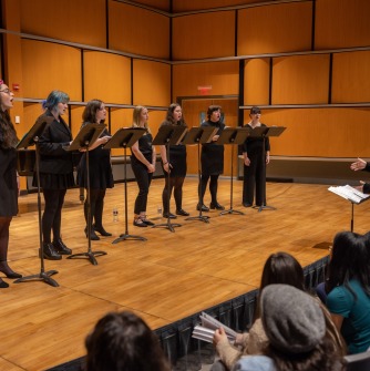 People standing on stage singing dressed in black clothing with music stands in front of them.
